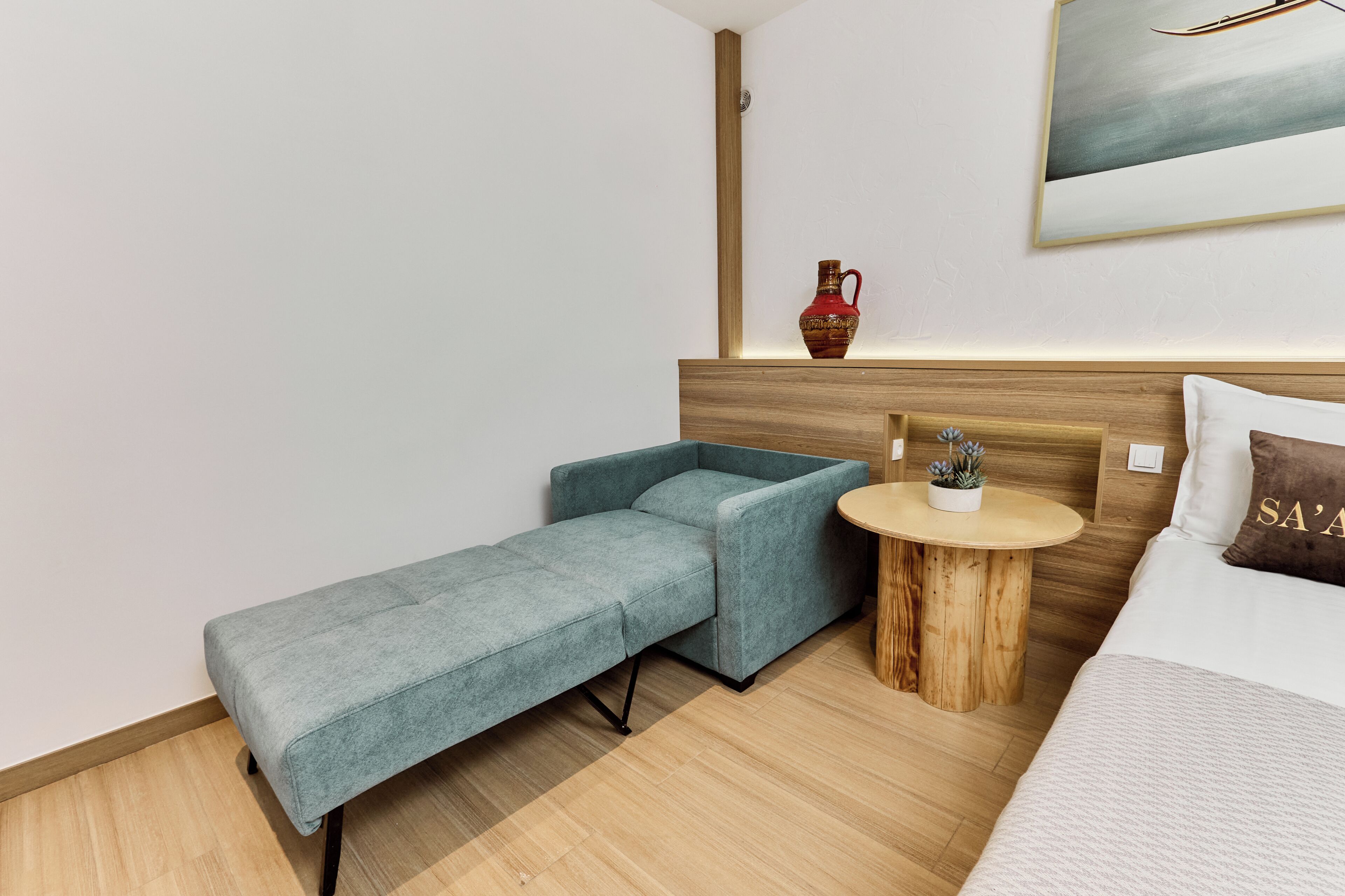 A modern hotel room with a bed, a small round wooden table, a blue sofa, and a decorative vase.