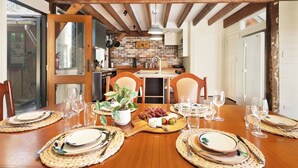 Comedor con mesa de madera dispuesta para una comida, cocina al fondo y pared de ladrillo.