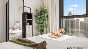 Et hotelværelse med en seng, et natbord med et bakke med mad, en potteplante, et spejl og et vindue med udsigt over byen.