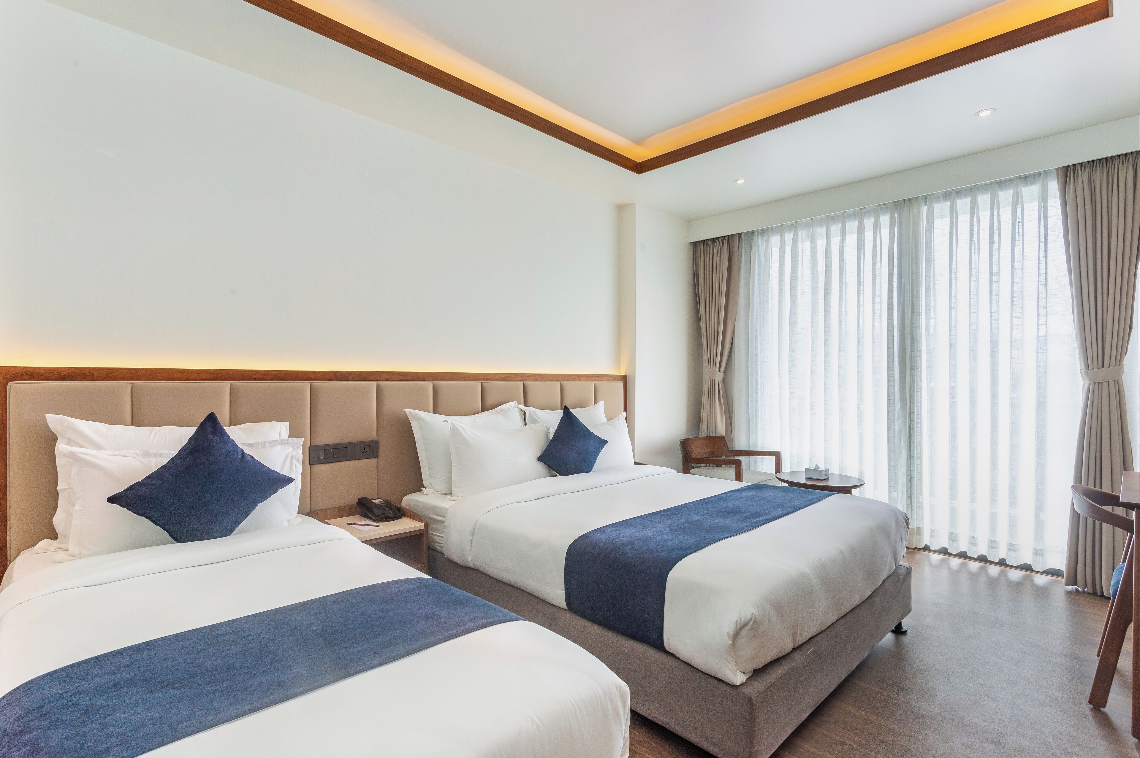 A hotel room with two beds, a wooden ceiling, a large window with curtains, and a bedside table with a lamp.