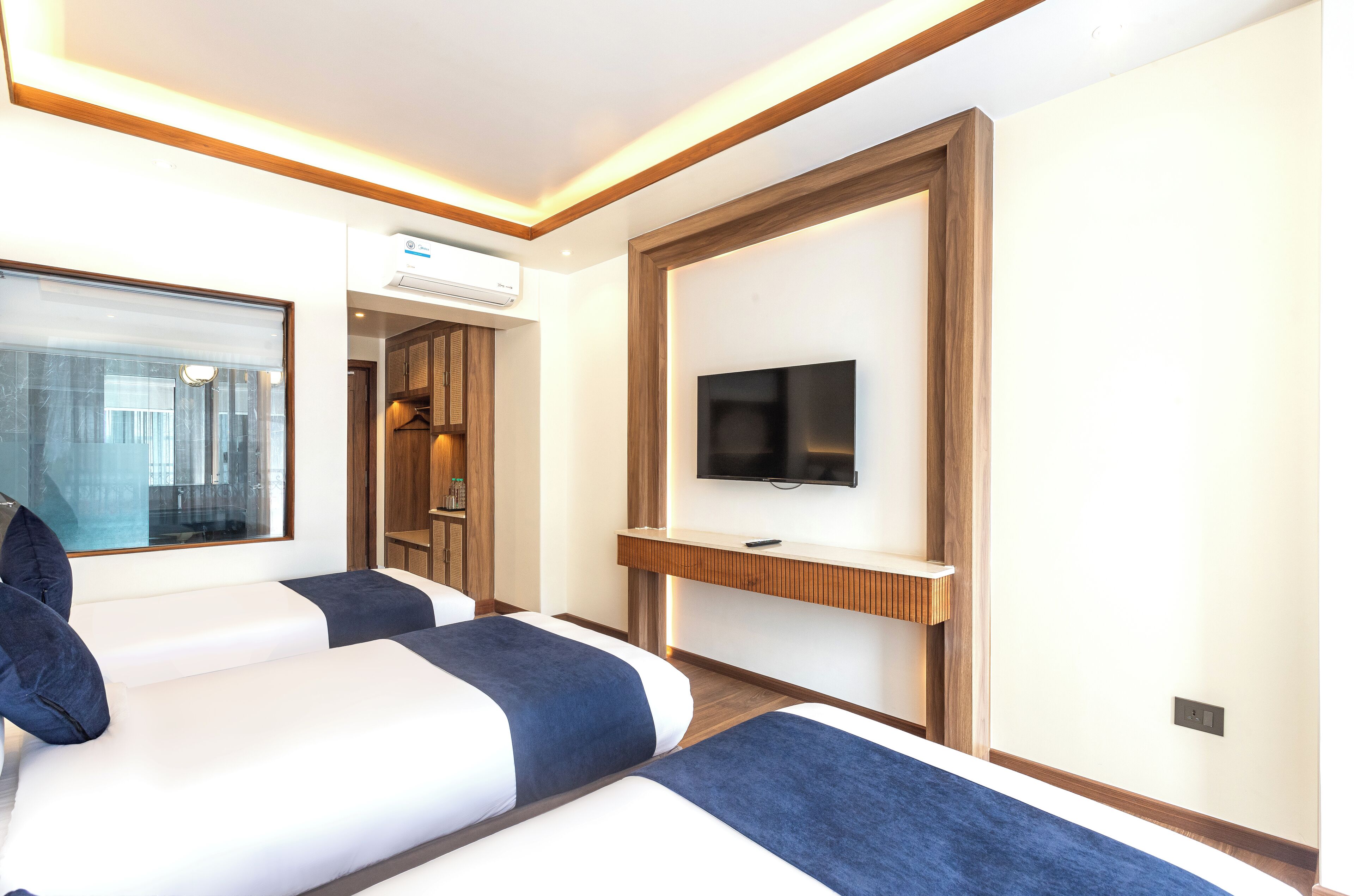 A hotel room with two beds, a TV mounted on a wooden panel, and an air conditioning unit on the wall.