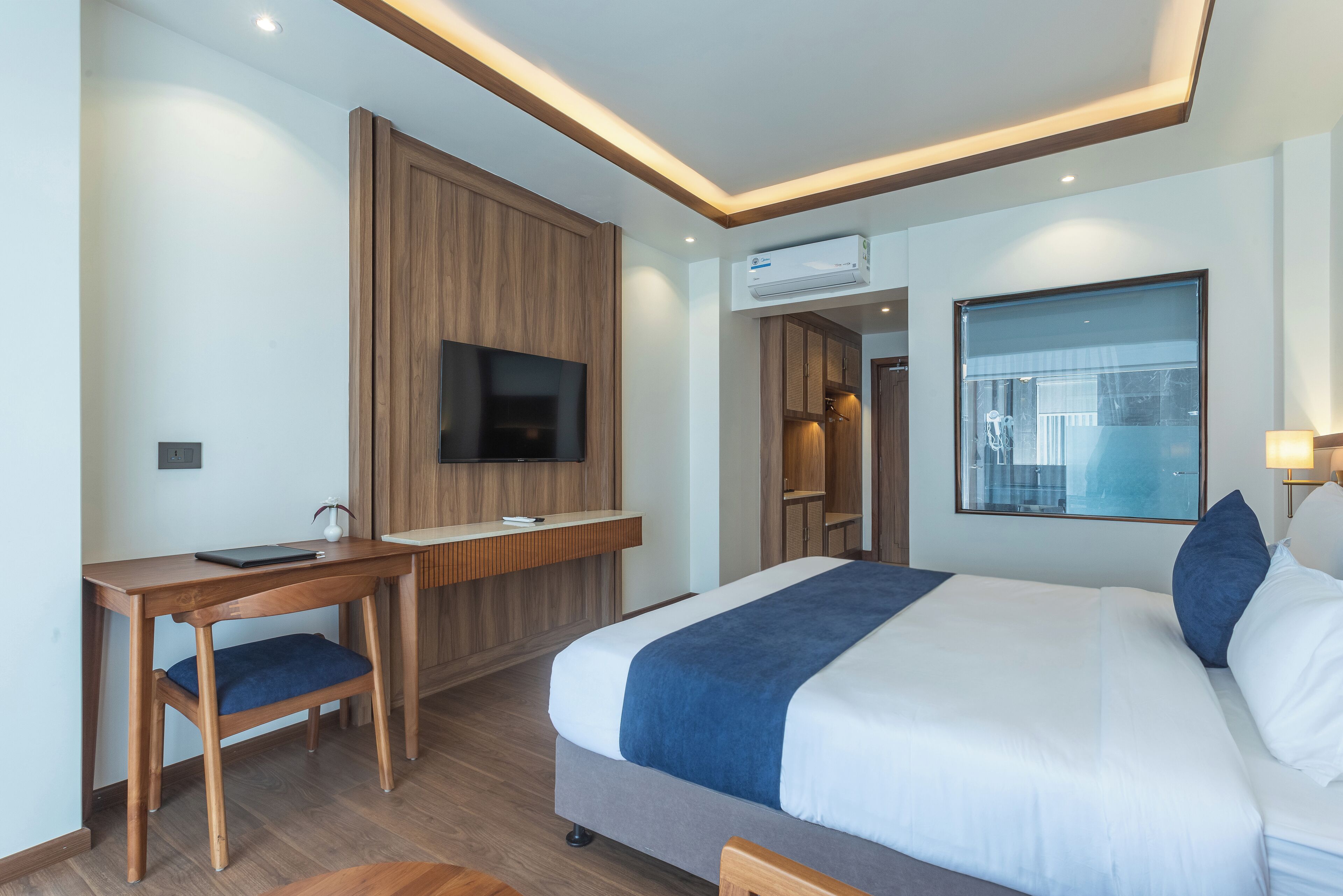 A modern hotel room with a large bed, a desk with a chair, a flat-screen TV, and an air conditioning unit.