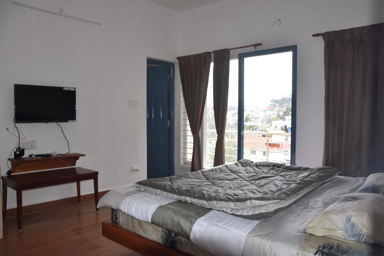 A bedroom with a bed, a TV mounted on the wall, a wooden bench, and a window with curtains.
