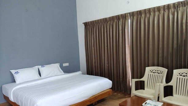 Habitación de hotel con cama de madera, ropa de cama blanca, mesita de noche de madera, una mesa con bandeja y dos sillas blancas.