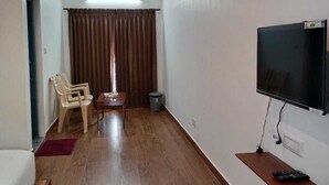 Una habitación con piso de madera, televisor montado en la pared, una silla, una mesita y una cortina.