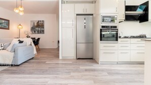A modern kitchen with white cabinetry, a built-in oven, and a refrigerator. There is a dining area with a table and chairs, and a sofa in the living room. - Modern Guesthouse with Private Pool • Close to Beaches, Shops & Restaurants • (Punaauia)