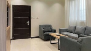 A modern living room with a dark wooden door, a grey sofa, a small table, and a window with sheer curtains. -  Arjan Park alkhalij (Riyadh)