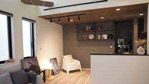 A modern living room with a sofa, a white armchair, a coffee table, and a TV. There is a kitchen area with a counter, shelves, and a clock.