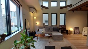 A modern living room with a sofa, a coffee table, and a large mirror. There are multiple windows providing natural light, and a plant is visible in the foreground.