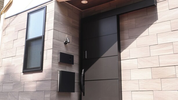 A modern building facade with a recessed entryway, a large window with a metal screen, and a door with a black handle.