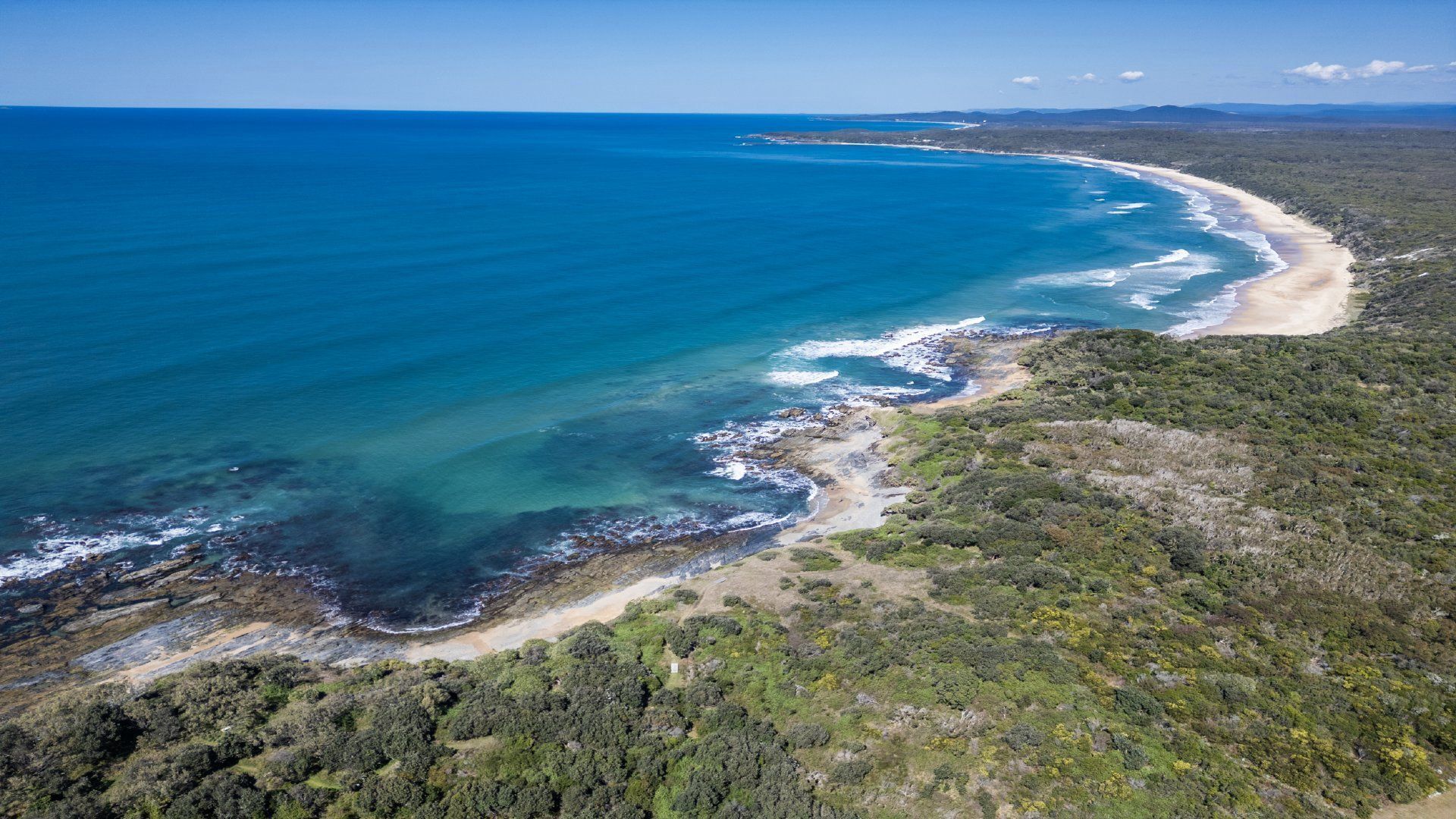 Vue aérienne d’une zone côtière avec une plage, une végétation luxuriante et des eaux d’un bleu limpide.