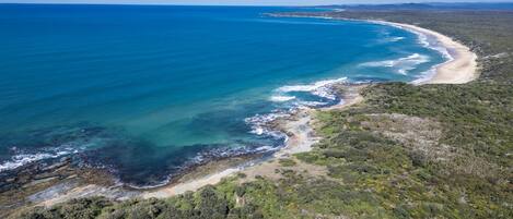 Vue aérienne d’une zone côtière avec une plage, une végétation luxuriante et des eaux d’un bleu limpide.