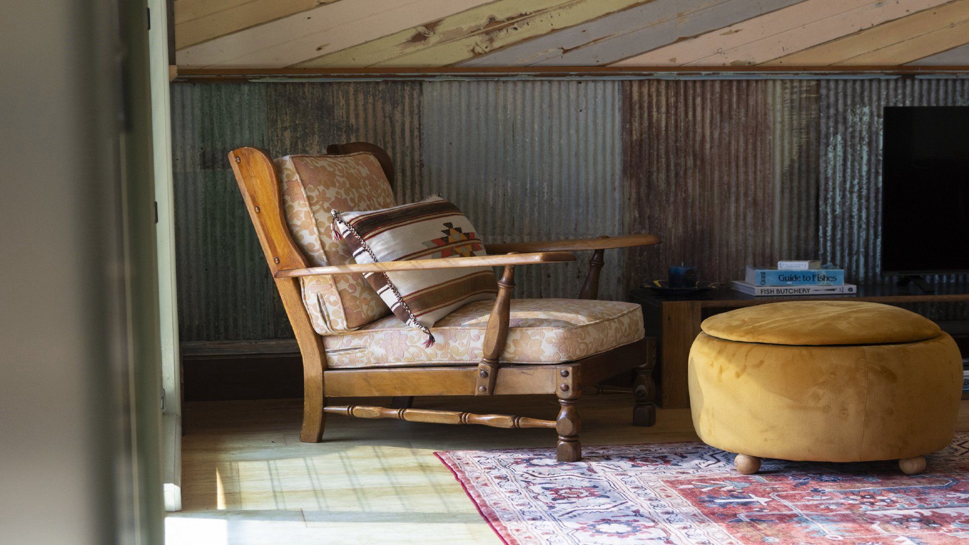 A room with a wooden chair, a small ottoman, a patterned rug, and a television against a corrugated metal wall.