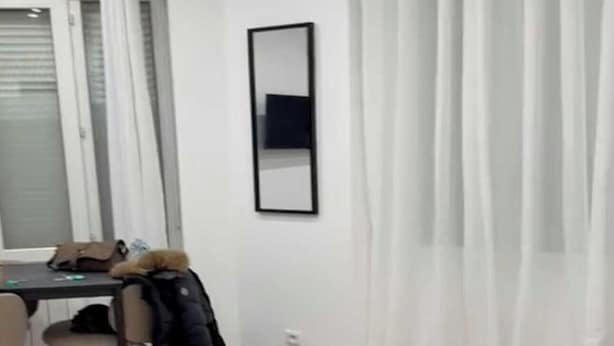 Ein kleiner, moderner Raum mit einem Esstisch, Stühlen, einem über einen Stuhl geworfenen Mantel, einem Spiegel an der Wand und durchsichtigen Vorhängen.
