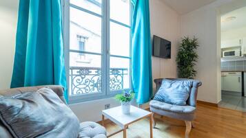 En moderne stue med sofa, lænestol og et lille bord med en plante. Et stort vindue med gardiner er synligt, og et køkkenområde ses gennem en åben dør.