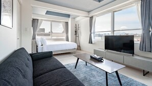 Una camera d'albergo moderna con un divano grigio, una TV a schermo piatto, un letto e ampie finestre con vista sulla città.