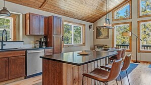Une cuisine avec des meubles en bois, un îlot central doté de tabourets de bar, de grandes fenêtres et un plafond en bois.