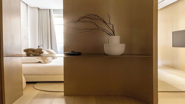 Une chambre moderne et minimaliste, avec un plancher en bois, un lit doté d’une tête de lit blanche, une table de chevet ornée d’un vase rempli de branches et un téléviseur fixé au mur.