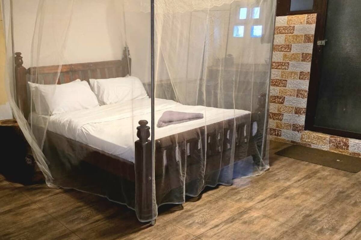 A room with a bed covered by a mosquito net, a wooden floor, a chair, and a wall-mounted fan.