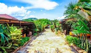 A stone pathway leading through a lush garden with tropical plants and a wooden pergola structure.