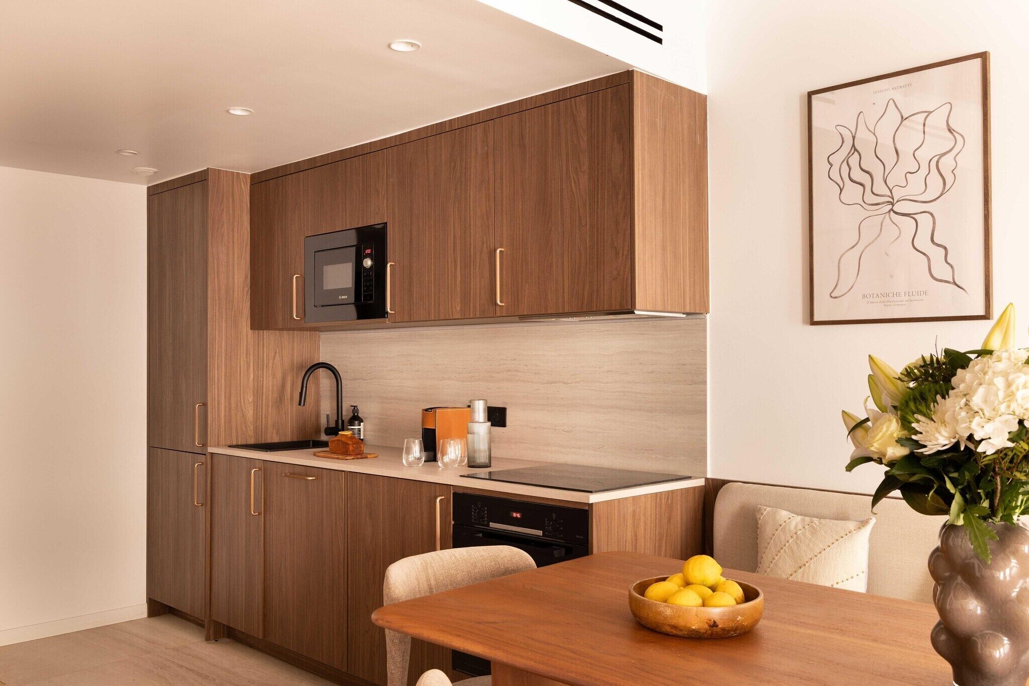 A modern kitchen with wooden cabinets, a black microwave, and a dining area with a wooden table and a bowl of lemons.