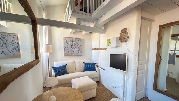 Una sala de estar moderna con un sofá, una otomana y una mesa redonda. Un televisor está montado en la pared, y hay obras de arte y una lámpara en la pared.
