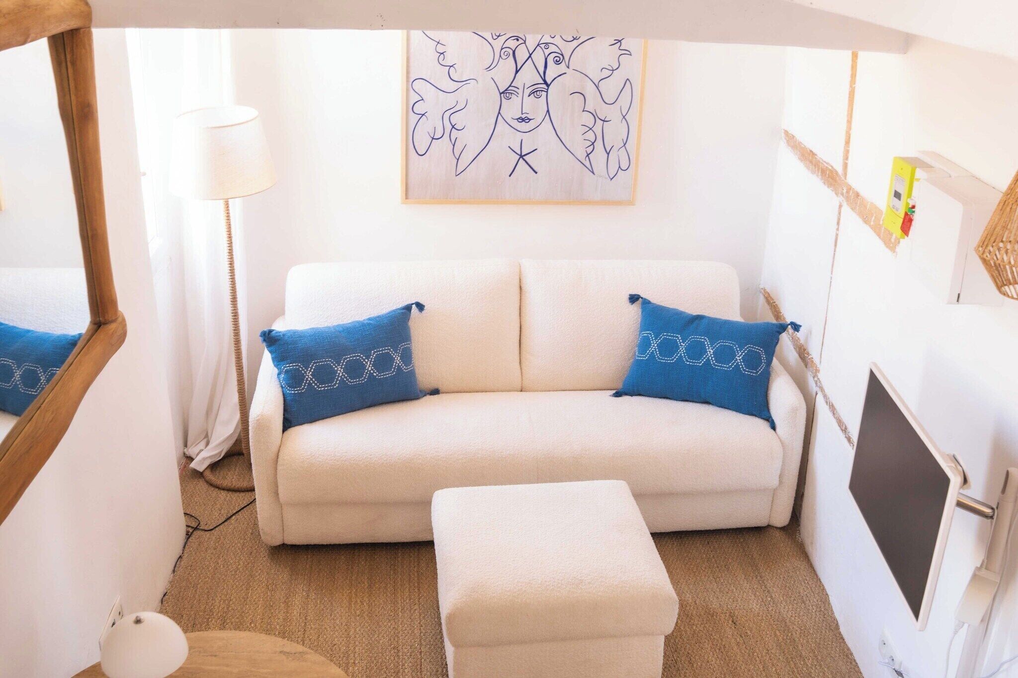 A cozy room with a white sofa, two blue pillows, a small ottoman, a wooden-framed mirror, a lamp, and a wall art piece.