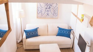 Una habitación acogedora con un sofá blanco, dos almohadas azules, un pequeño otoman, un espejo con marco de madera, una lámpara y una obra de arte en la pared.