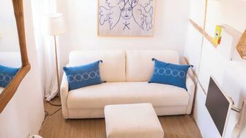 A cozy room with a white sofa, two blue pillows, a small ottoman, a wooden-framed mirror, a lamp, and a wall art piece.