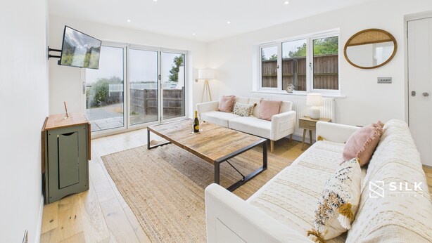 A spacious living room with a large sliding glass door leading to a balcony, a white sofa set, a wooden coffee table, and a television mounted on the wall.