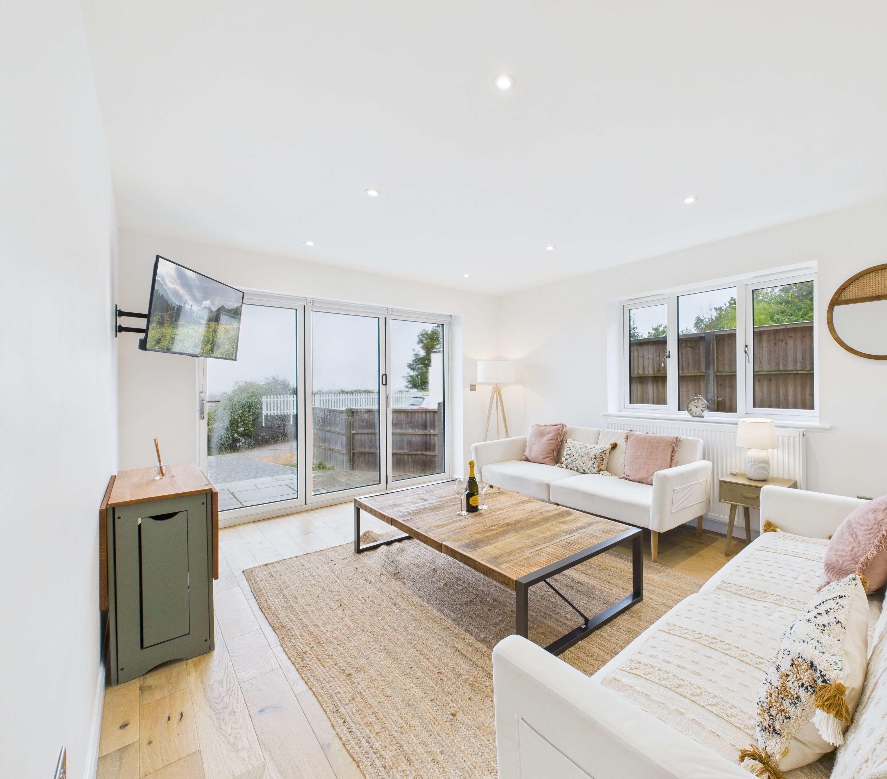 A spacious living room with a large sliding glass door leading to a balcony, a white sofa set, a wooden coffee table, and a television mounted on the wall.