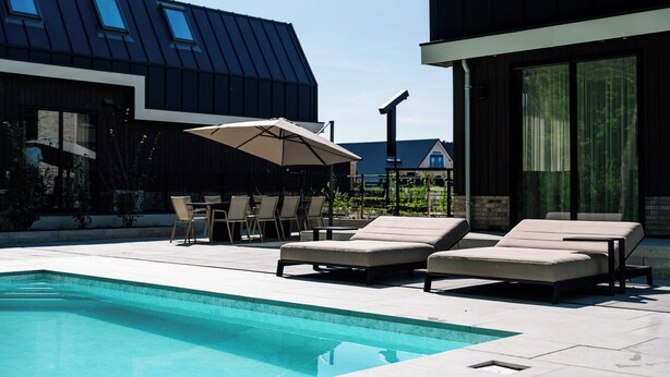 A modern outdoor pool area with lounge chairs and a table under a large umbrella, surrounded by buildings with large windows and a sloped roof.
