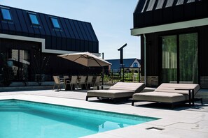 Een modern buitenzwembad met ligbedden en een tafel onder een grote parasol, omringd door gebouwen met grote ramen en een schuin dak.