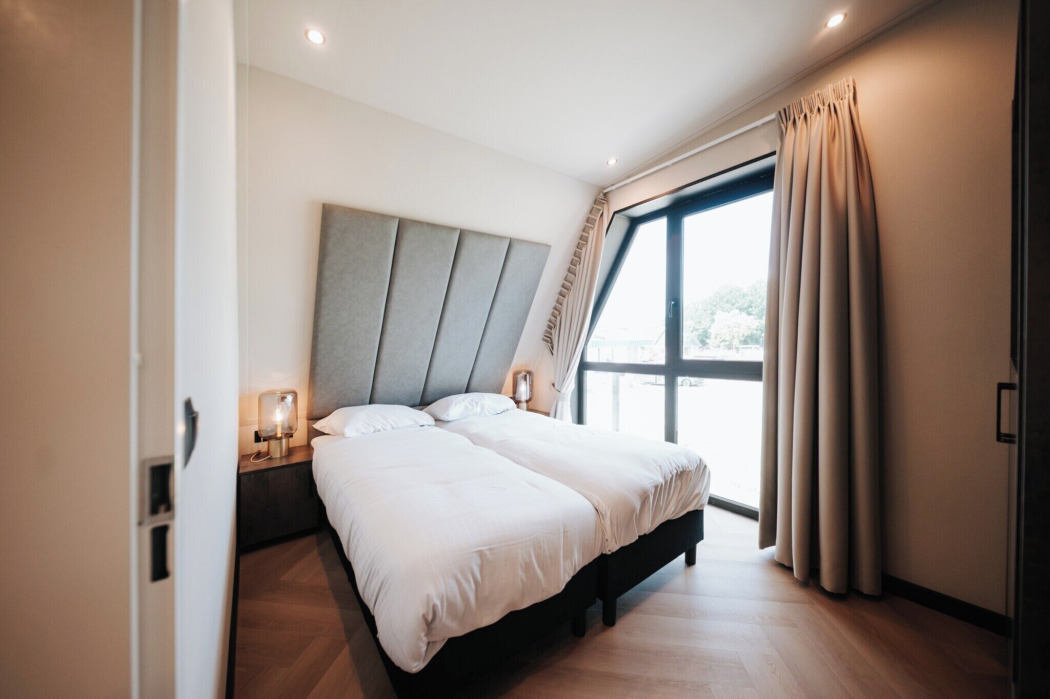 A modern bedroom with a large bed, a bedside table with a lamp, and a view of the outdoors through a large window.