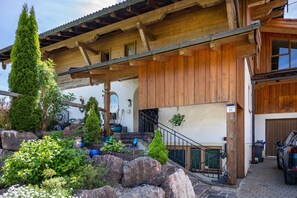 Ein Holzhaus mit Steinstufen, ein geparktes Auto und ein Garten mit verschiedenen Pflanzen und Steinen.