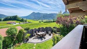 Une terrasse en bois avec des meubles d’extérieur, un mur en pierre et une vue sur une vallée verdoyante, avec des montagnes en arrière-plan.