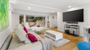 A modern living room with a white sofa, a coffee table, and a TV. There is a large window with a view of a garden and a hammock outside.
