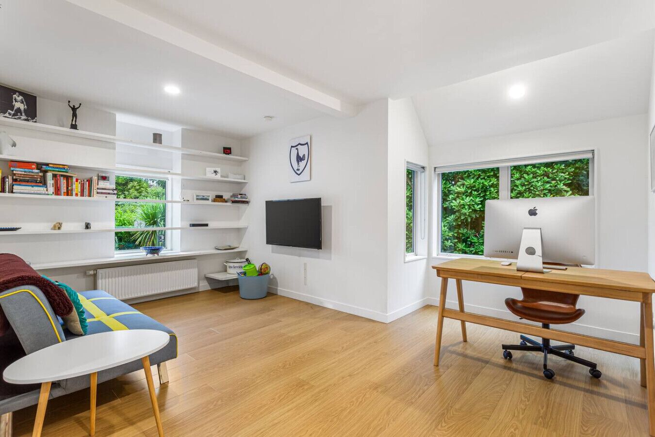 Ein modernes Wohnzimmer mit Holzboden, einem grauen Sofa, einem weißen Couchtisch, einem Holzbüro mit Computer, einem Bücherregal und einem Fernseher an der Wand.