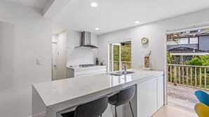 A modern kitchen with a white countertop, two black bar stools, a stainless steel range hood, and a view of a balcony with a wooden railing.