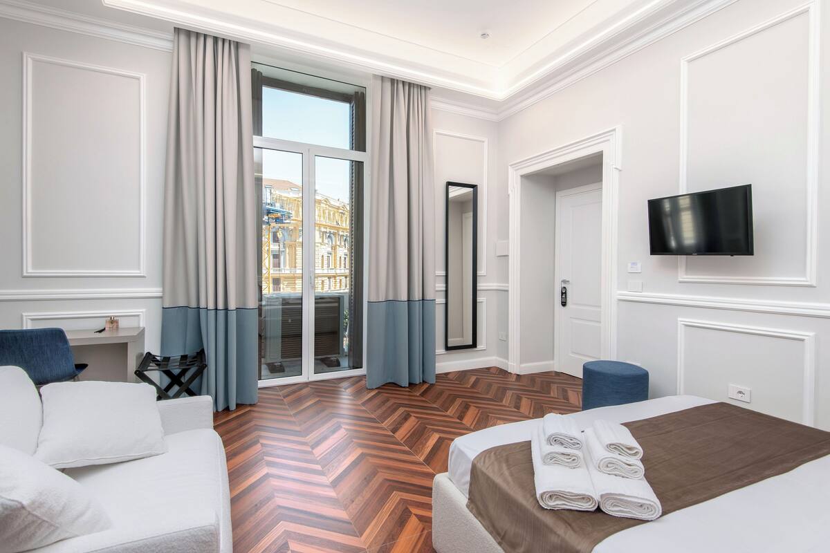 A hotel room with a large bed, a sofa, a television, and a view of a cityscape through the window.