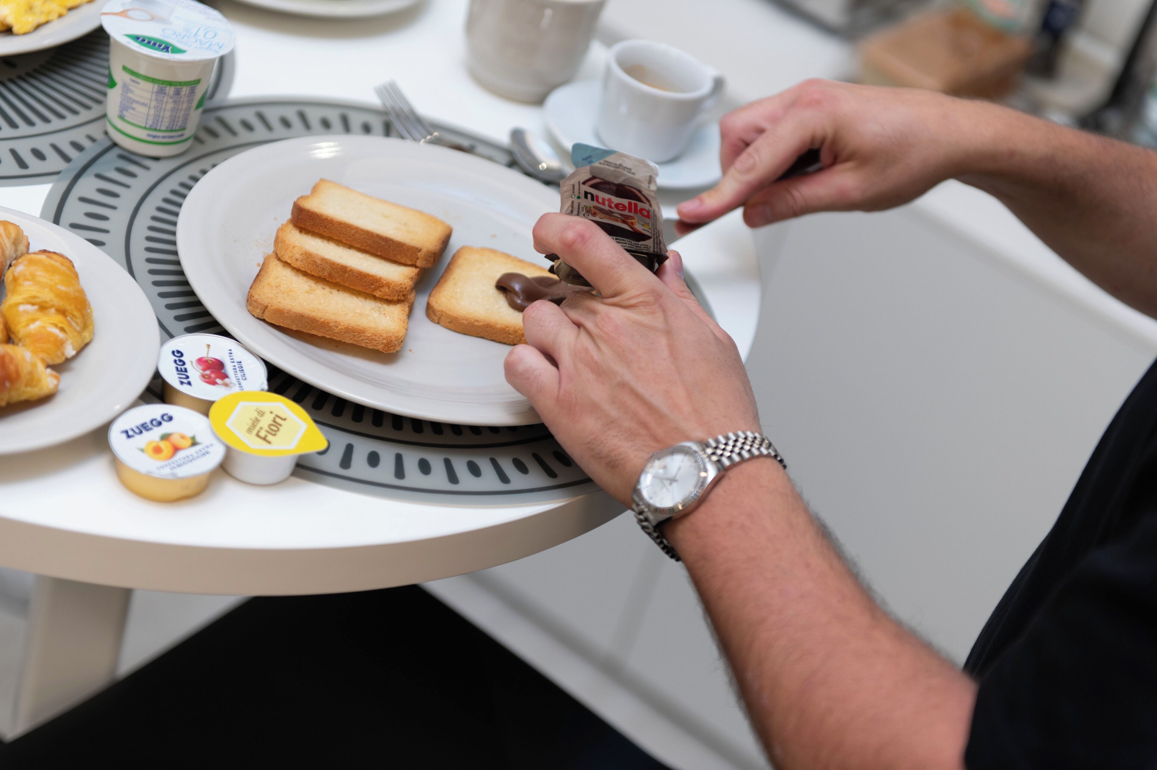 Una persona spalma Nutella su una fetta di pane a tavola, con croissant, formaggio e yogurt.