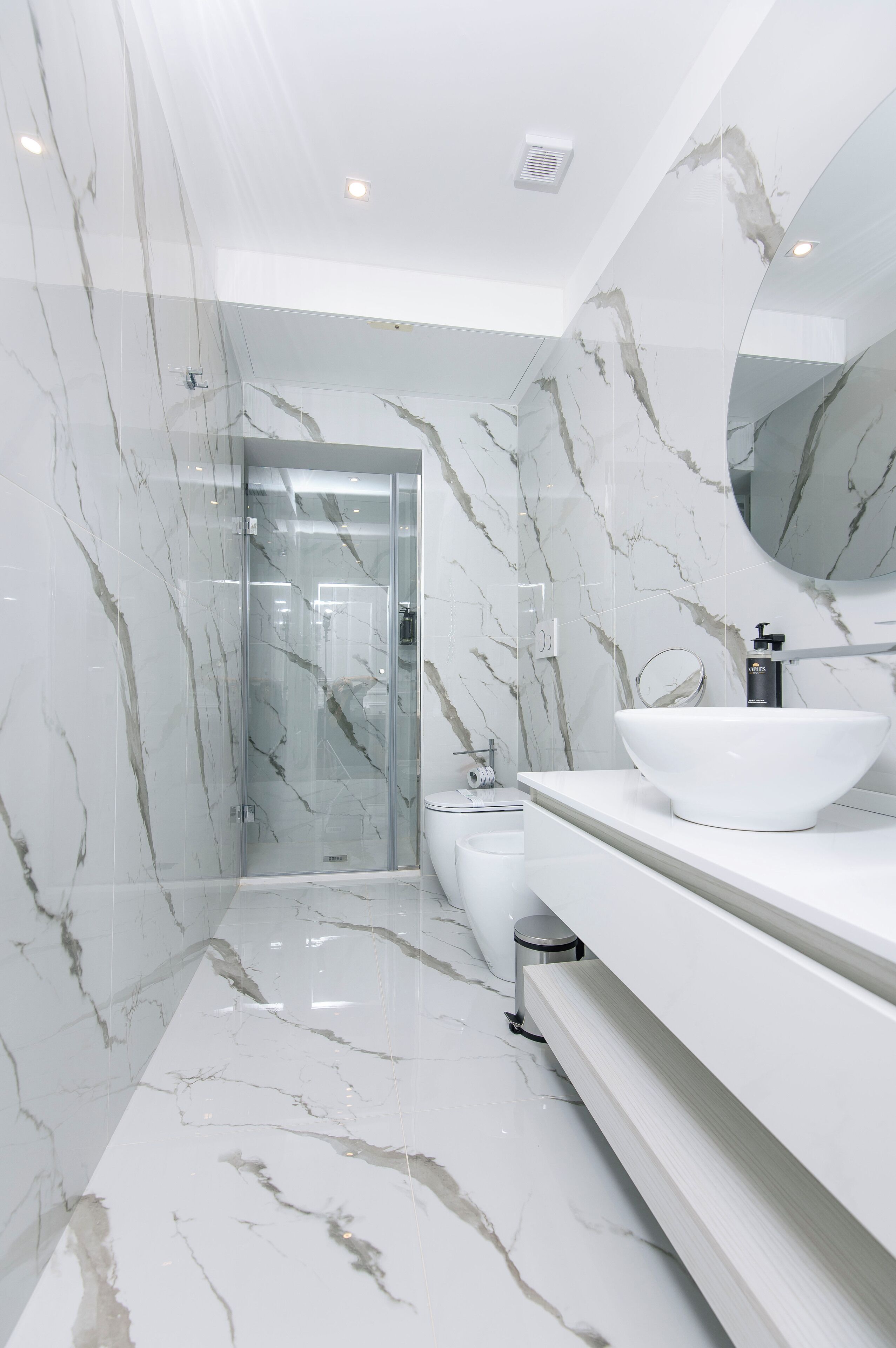 Une salle de bain moderne avec des murs en marbre, une douche en verre, un lavabo blanc et un grand miroir.