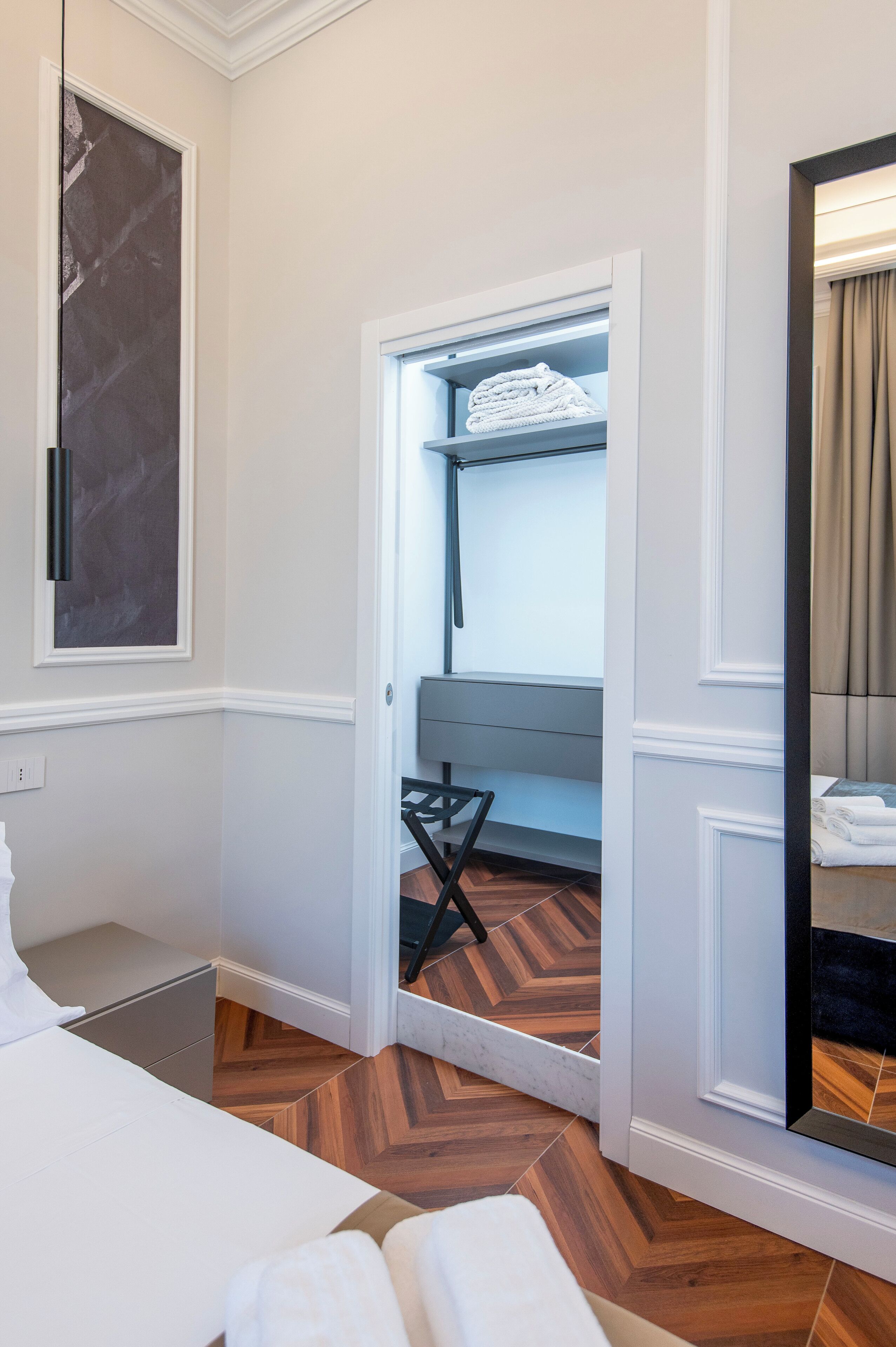 A hotel room with a wooden herringbone floor, a bed with white linens, a small seating area with a bench, and a wardrobe with a mirror.