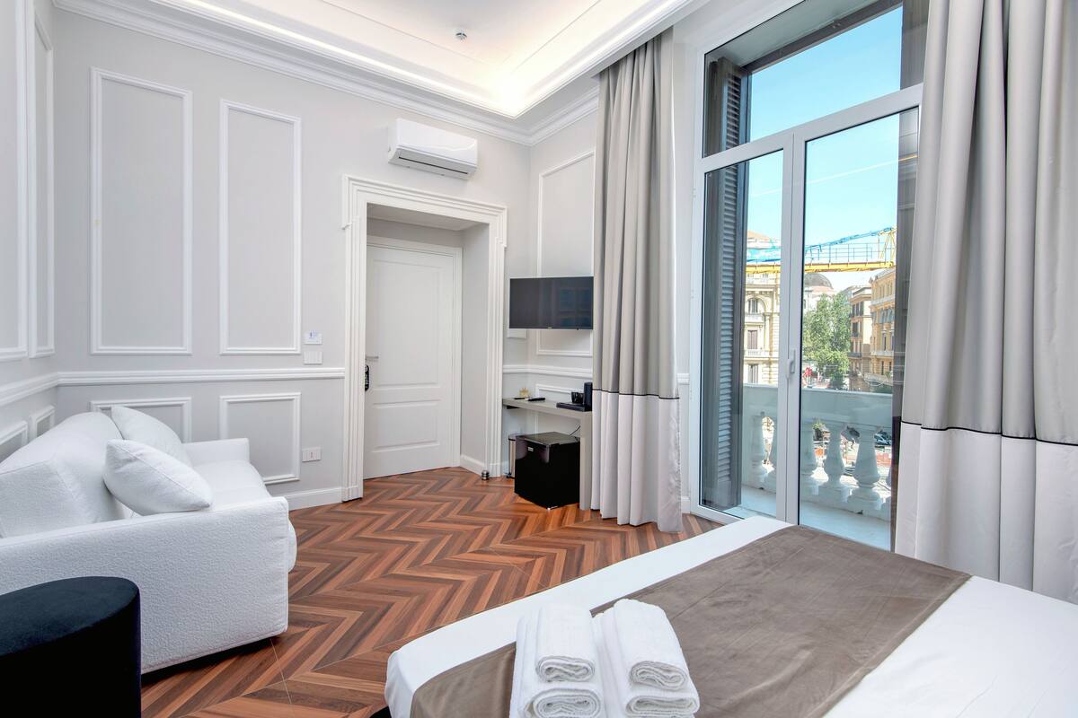 A hotel room with a bed, a sofa, a TV, and a balcony with a view of buildings.
