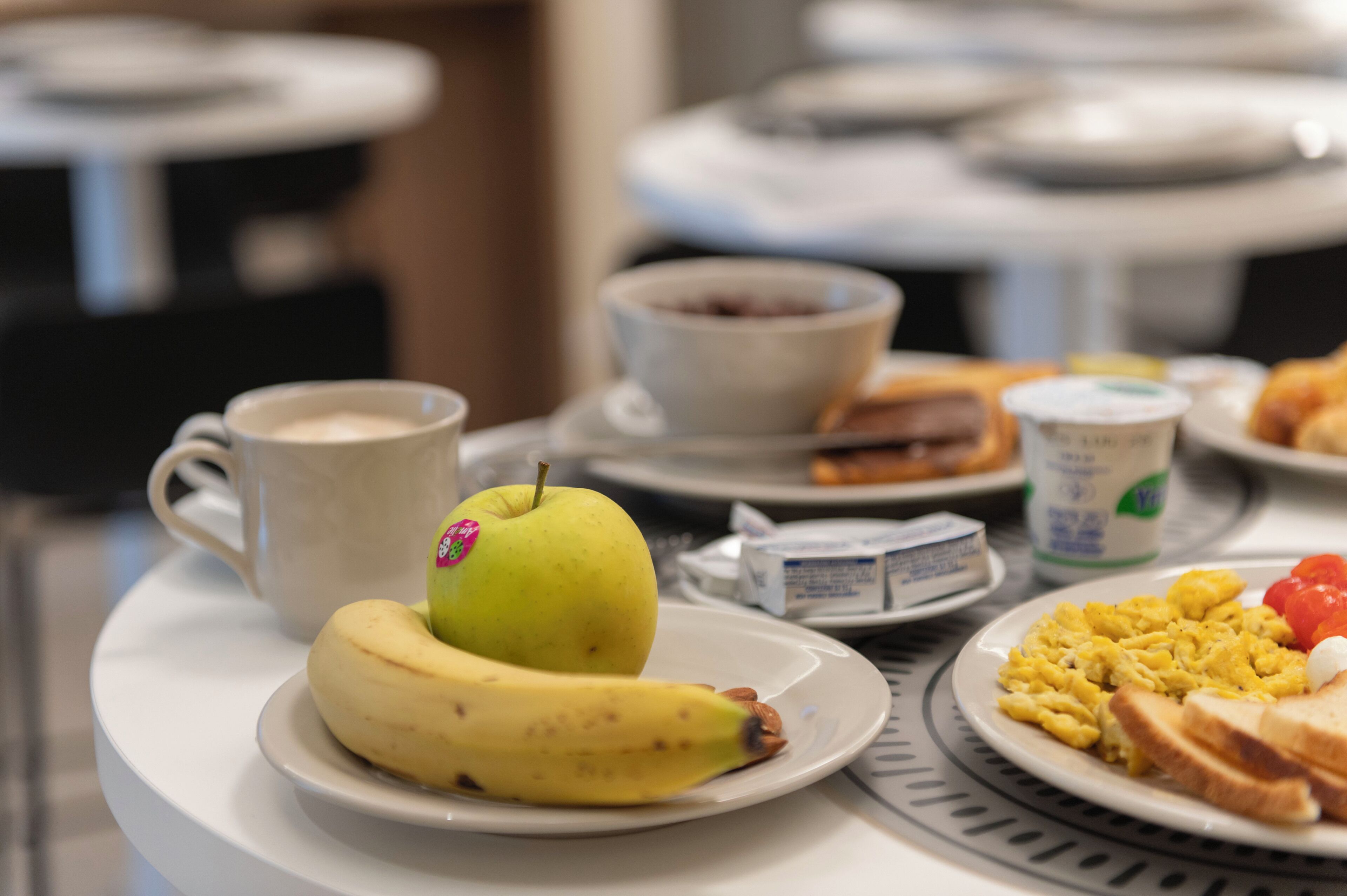 Une table de petit-déjeuner avec une assiette d’œufs brouillés, des toasts, une banane, une pomme, une tasse de café et un bol de céréales.
