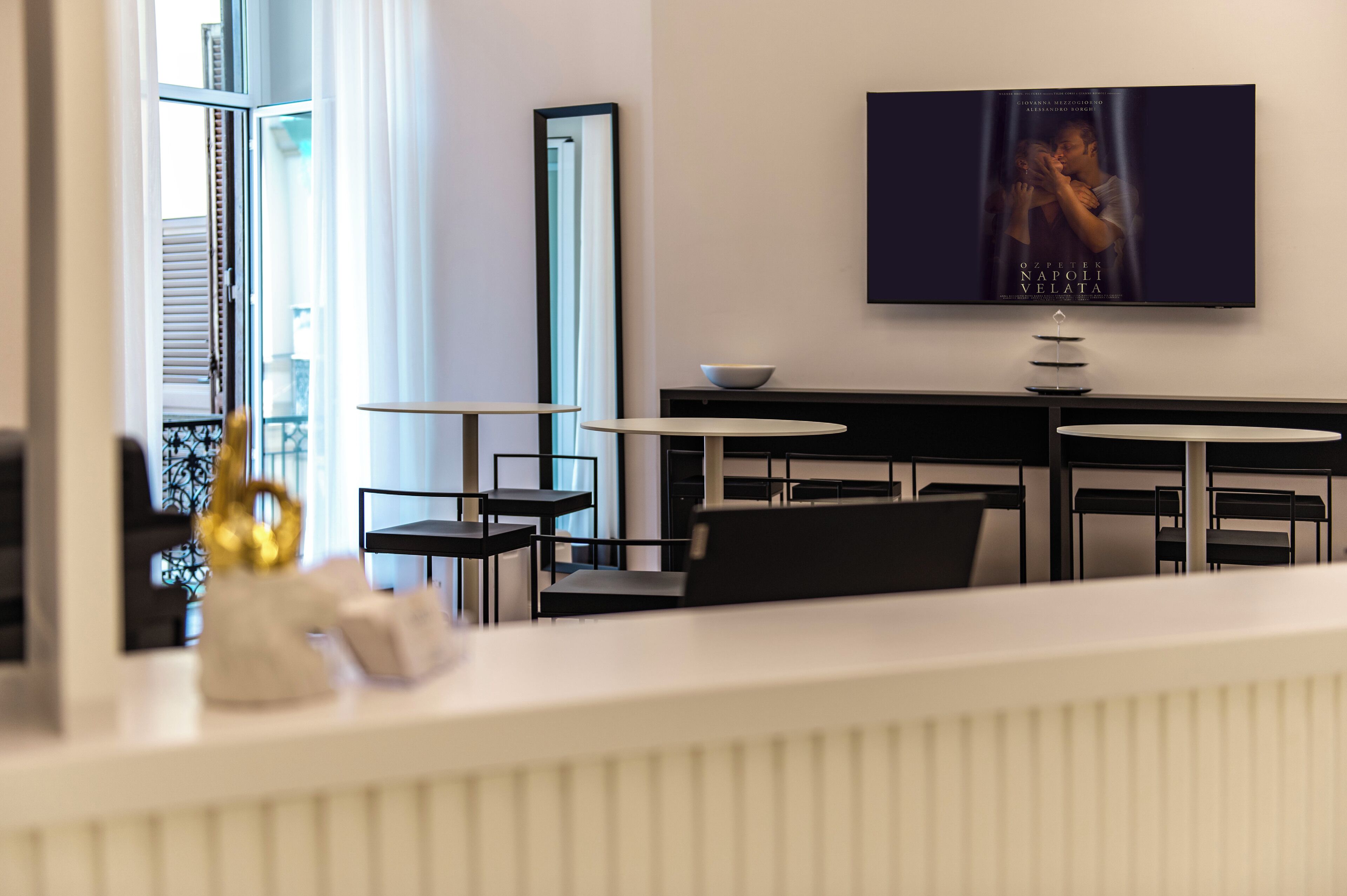 Una habitación de hotel moderna con una televisión de pantalla plana montada en la pared, un rincón de comedor con una mesa y sillas blancas, y un ventanal con persianas.