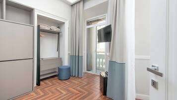 Ett modernt hotellrum med ett fiskbensmönstrat trägolv, en inbyggd garderob, en sittgrupp med en blå pall och en skjutdörr i glas som leder ut till en balkong.