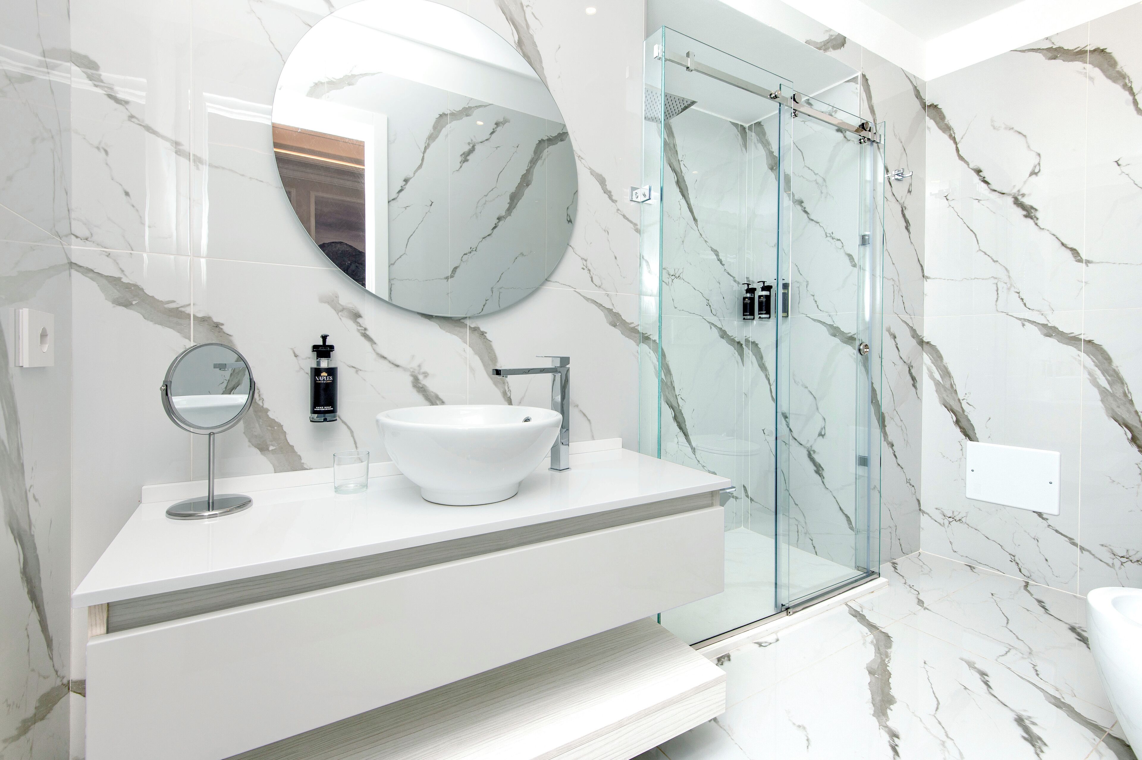 A modern bathroom with a white marble vanity, a round mirror, a vessel sink, and a glass-enclosed shower.