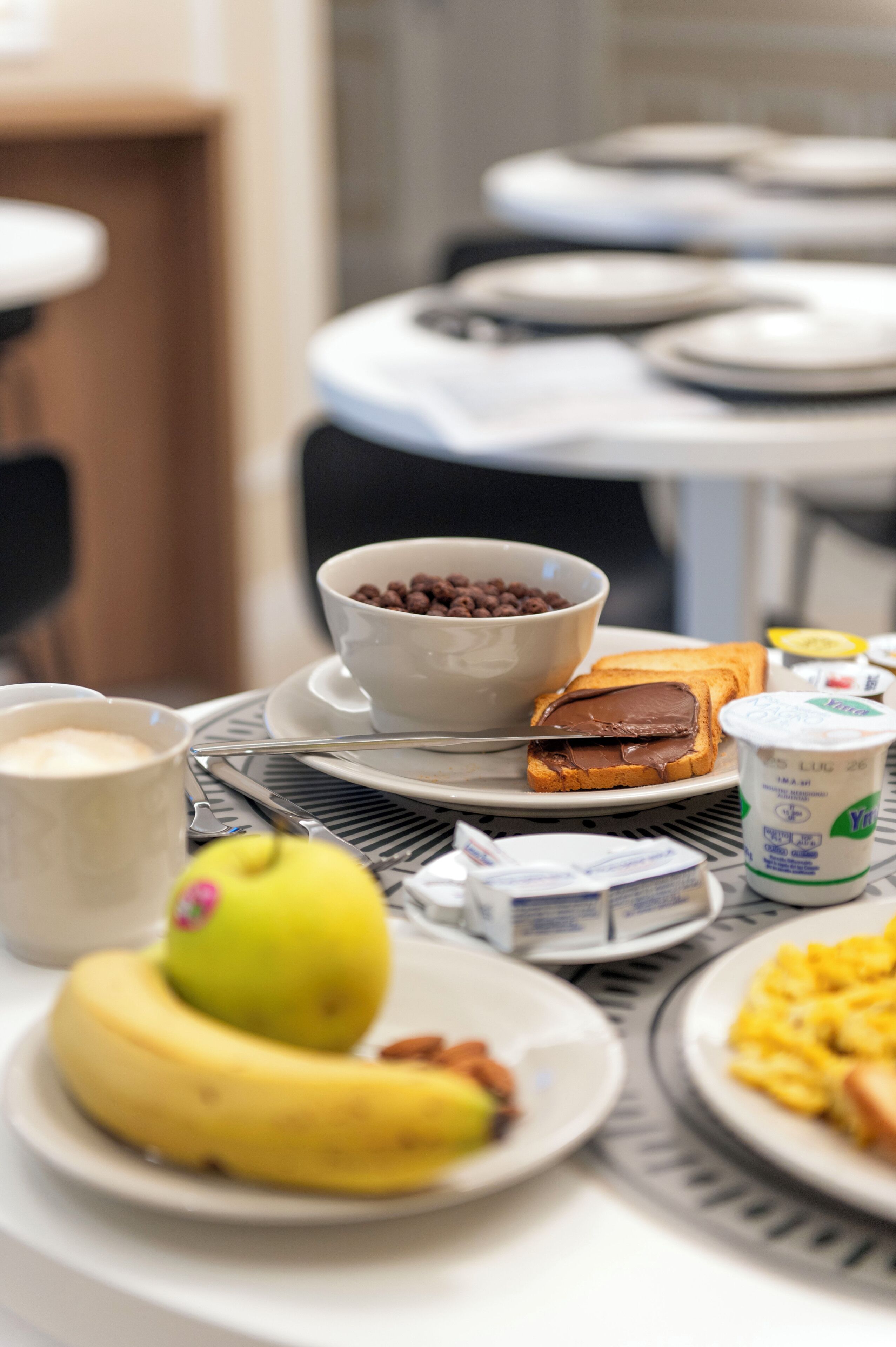 Una mesa de desayuno con un tazón de cereal, tostadas con crema de chocolate, un plátano, una taza de café y un plato de huevos revueltos.