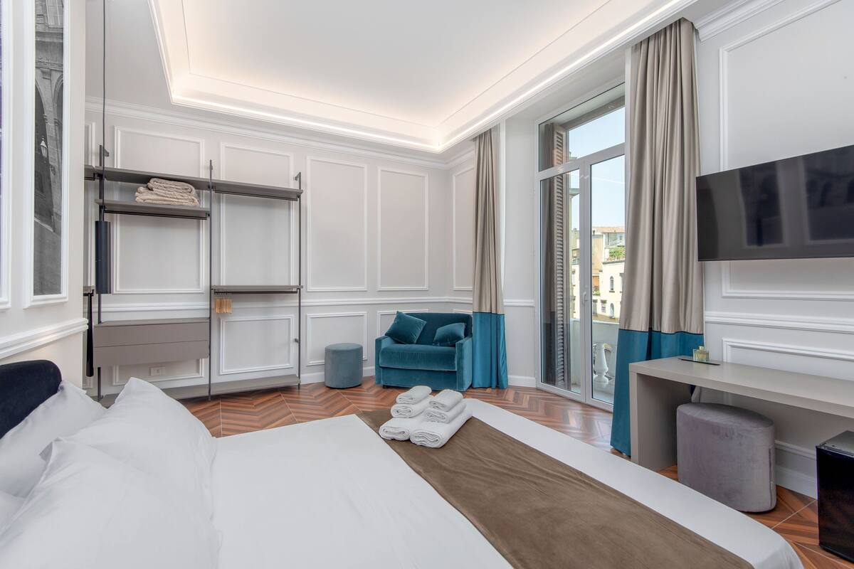 A hotel room with a bed, a sofa, a TV, a desk, and a window with curtains.